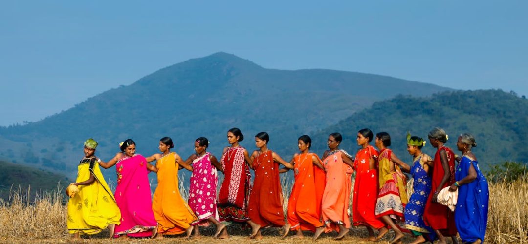 A group of women perform a traditional tribal dance in vibrant clothing against a rural Indian backdrop.