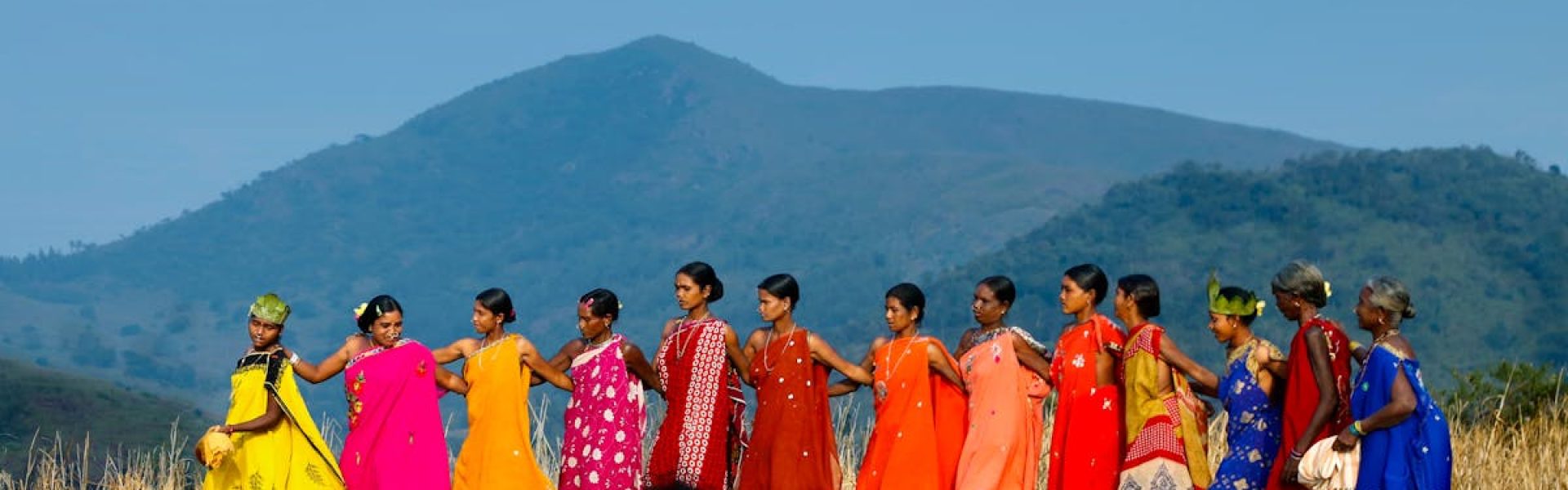 A group of women perform a traditional tribal dance in vibrant clothing against a rural Indian backdrop.